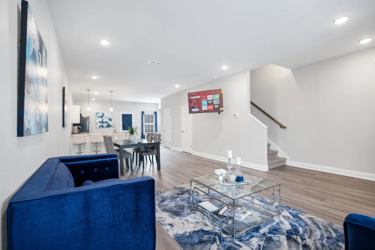 The image showcases a bright, open-concept living area featuring a plush blue sofa, sleek glass coffee table, large television, and a stylish dining area. The space is modern and inviting, perfect for relaxing, entertaining, or gathering for meals. Ample natural light and contemporary décor create a warm, welcoming atmosphere.