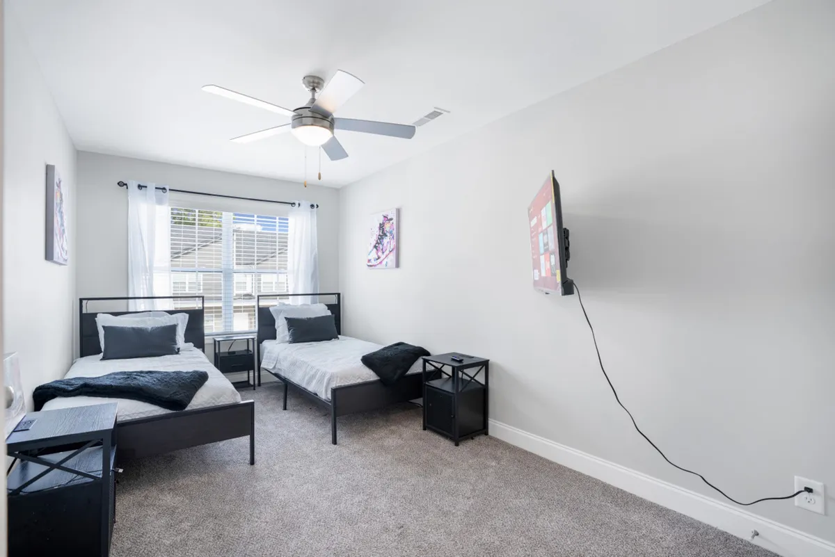 A spacious bedroom designed for flexibility, featuring two twin beds, a large window letting in ample light, a ceiling fan, and a wall-mounted TV. The room is styled for comfort, making it ideal for kids, friends, or family to share sleepovers or movie nights together.