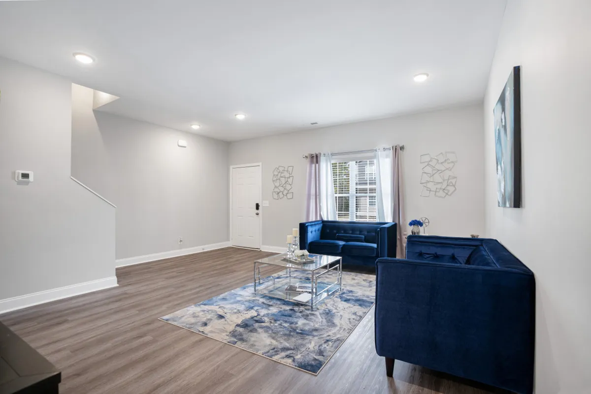 A bright, open-concept living area with blue sofas, a glass coffee table, wall art, and a large window letting in natural light. The space is inviting and versatile, suitable for relaxing, socializing, or setting up a remote workspace.