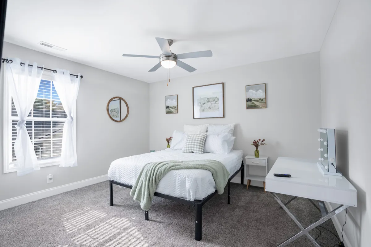 A tranquil bedroom featuring a bed with crisp white bedding, decorative wall art above the headboard, a window draped with sheer curtains letting in morning light, and a modern desk ideal for reading or working. The room exudes a peaceful, restful atmosphere, enhanced by soft textiles and muted colors.