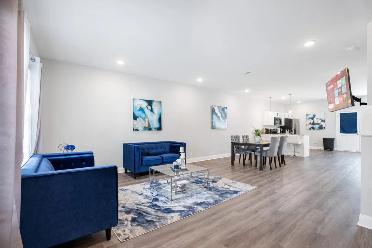 The shot features plush blue sofas and abstract wall art, with a dining table and chairs in the background, all unified by a soft area rug. Natural light pours in through unseen windows, highlighting the comfortable, contemporary furnishings throughout the open layout.