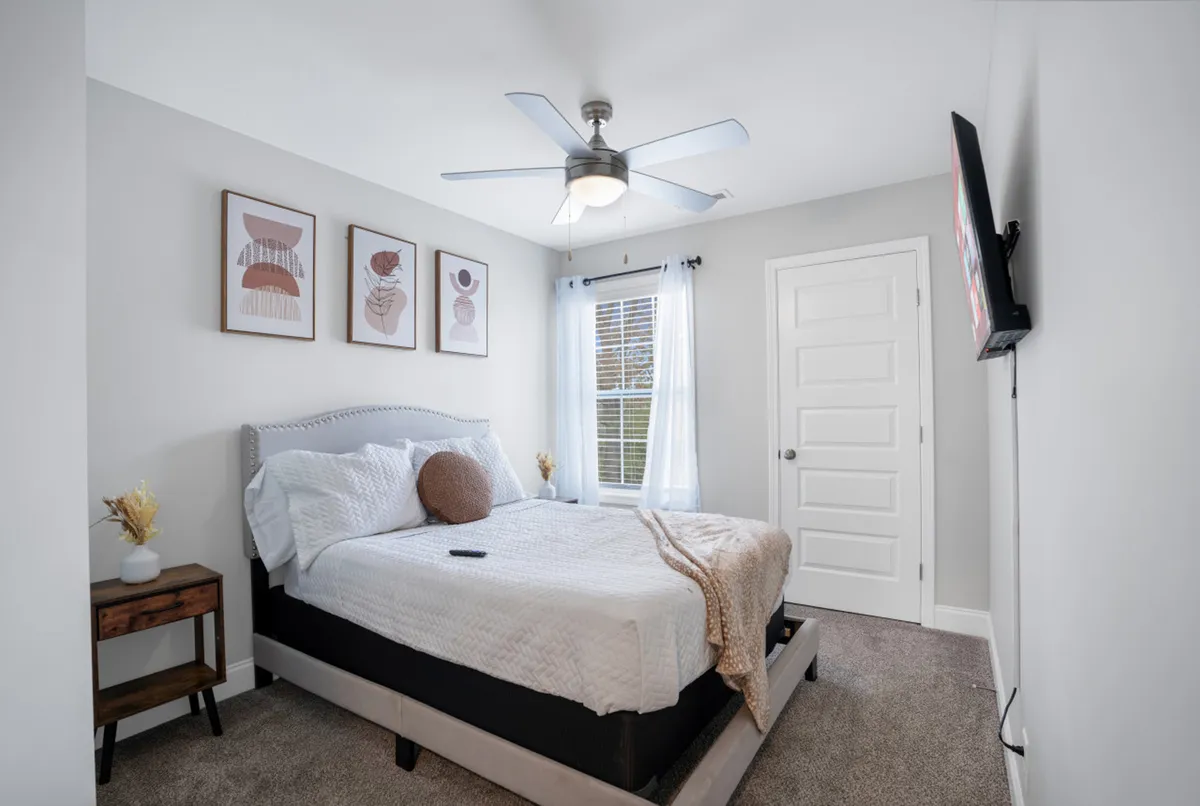 A cozy bedroom with a bed adorned with decorative pillows and soft blankets, complemented by wall art and a nightstand with artistic décor. The sunlight filters through the window, creating a warm and inviting environment ideal for relaxing and unwinding.